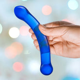 A hand elegantly grips the Gläs 6" Curved G-Spot Blue Glass Dildo, masterfully designed for optimal G-Spot stimulation. The softly blurred background shimmers with multi-colored bokeh lights, enhancing the allure of this captivating product from Gläs.